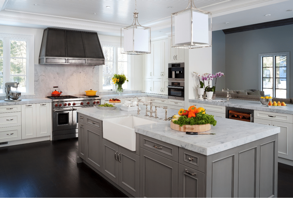 Remodeled kitchen with dark wood floors, large gray island with white stone countertop and white farmhouse sink. White cabinets, stainless-steel stove, and pendant lighting over island.