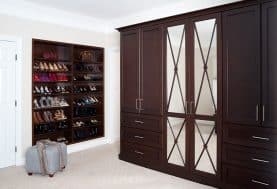 A closet with white walls and carpet accented with dark wood closet doors and shoe storage and a gray ottoman