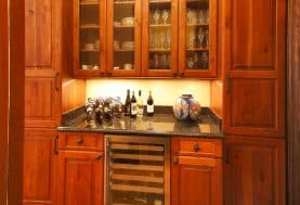 Warm-toned cabinetry with glass doors displaying glassware and a black countertop displaying wine above a wine fridge