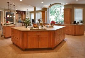 A kitchen with a large window with beige counter tops and warm wooden cabinetry and beige tiles.