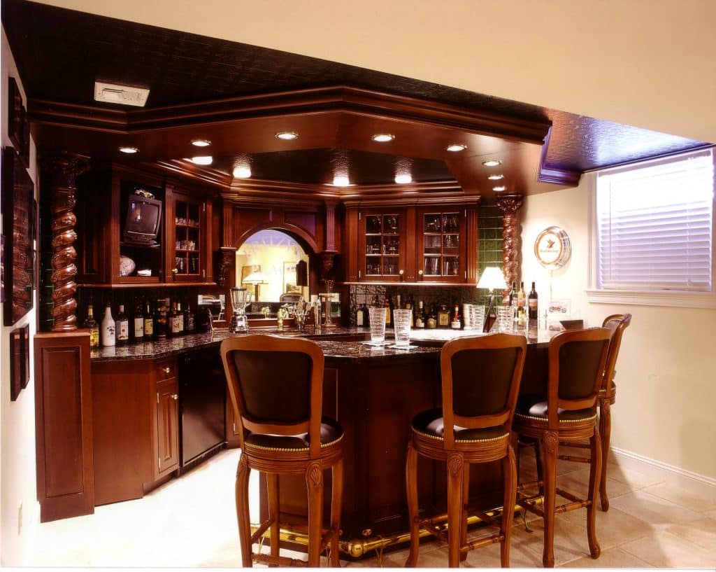 Remodeled wet bar in a basement, with light floors, dark wood shelving, cabinetry, and island, dark granite countertops, and dark wood stools at the island.