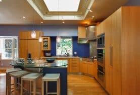 A modern kitchen with a dark blue backsplash, warm wood cabinetry, a skylight and a rounded island with a glass countertop.