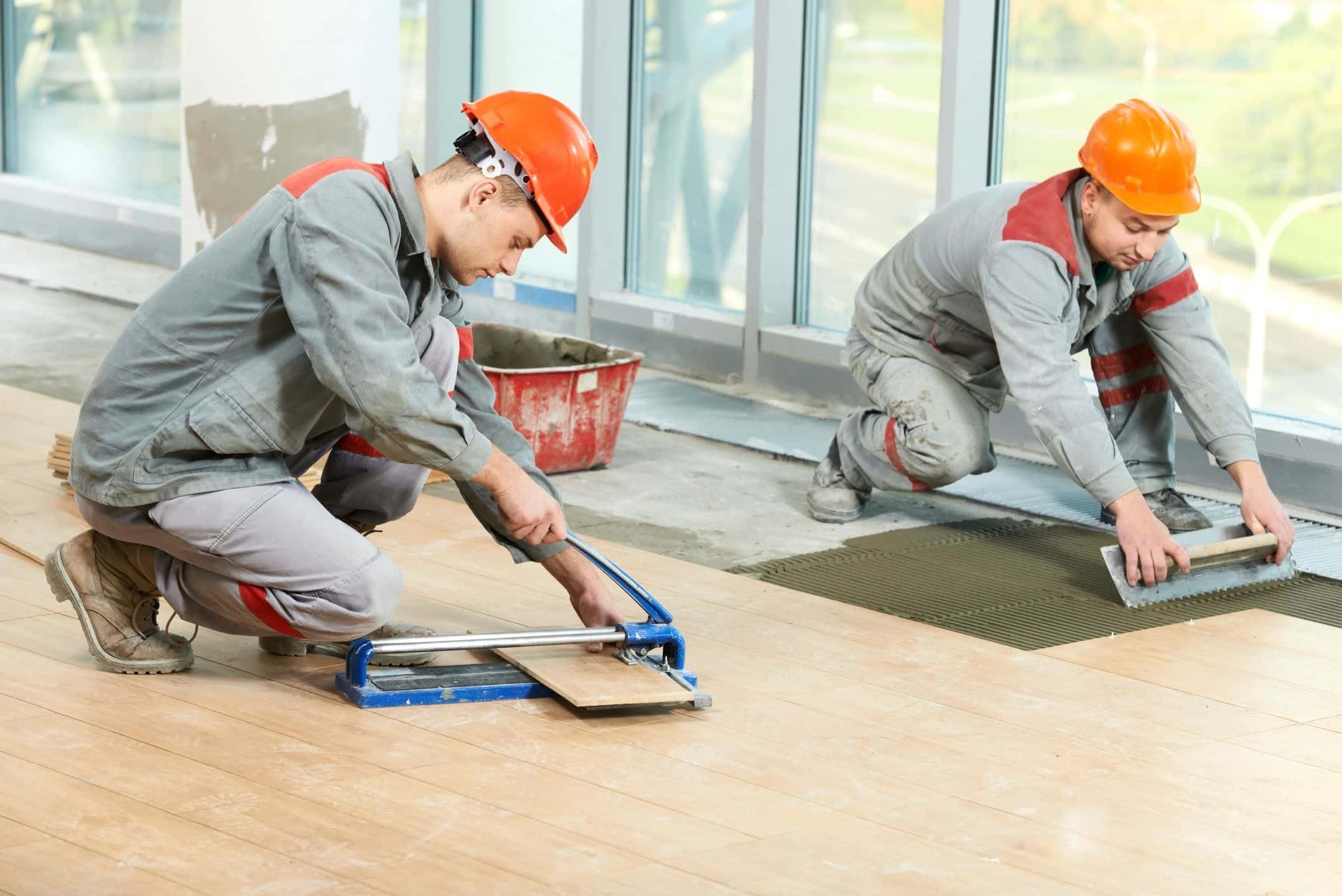 Technicians laying wood flooring panels on a floor, while wearing orange hard hats and gray uniforms.