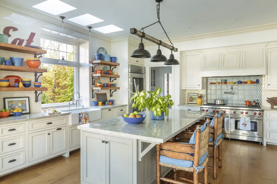 White kitchen with large island and open shelving. Light wood floors, large window over the sink, white stone countertops, and double gas stoves.